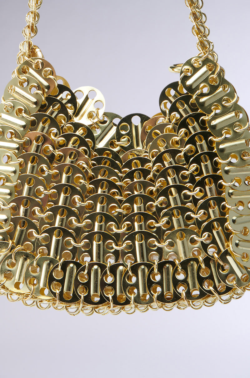 A close-up of the TAYTAY METAL PURSE, a gold metallic mini baguette bag crafted from interlocking metal pieces with curved and rectangular links, giving it a textured, shiny surface against a plain, light-grey background. 9