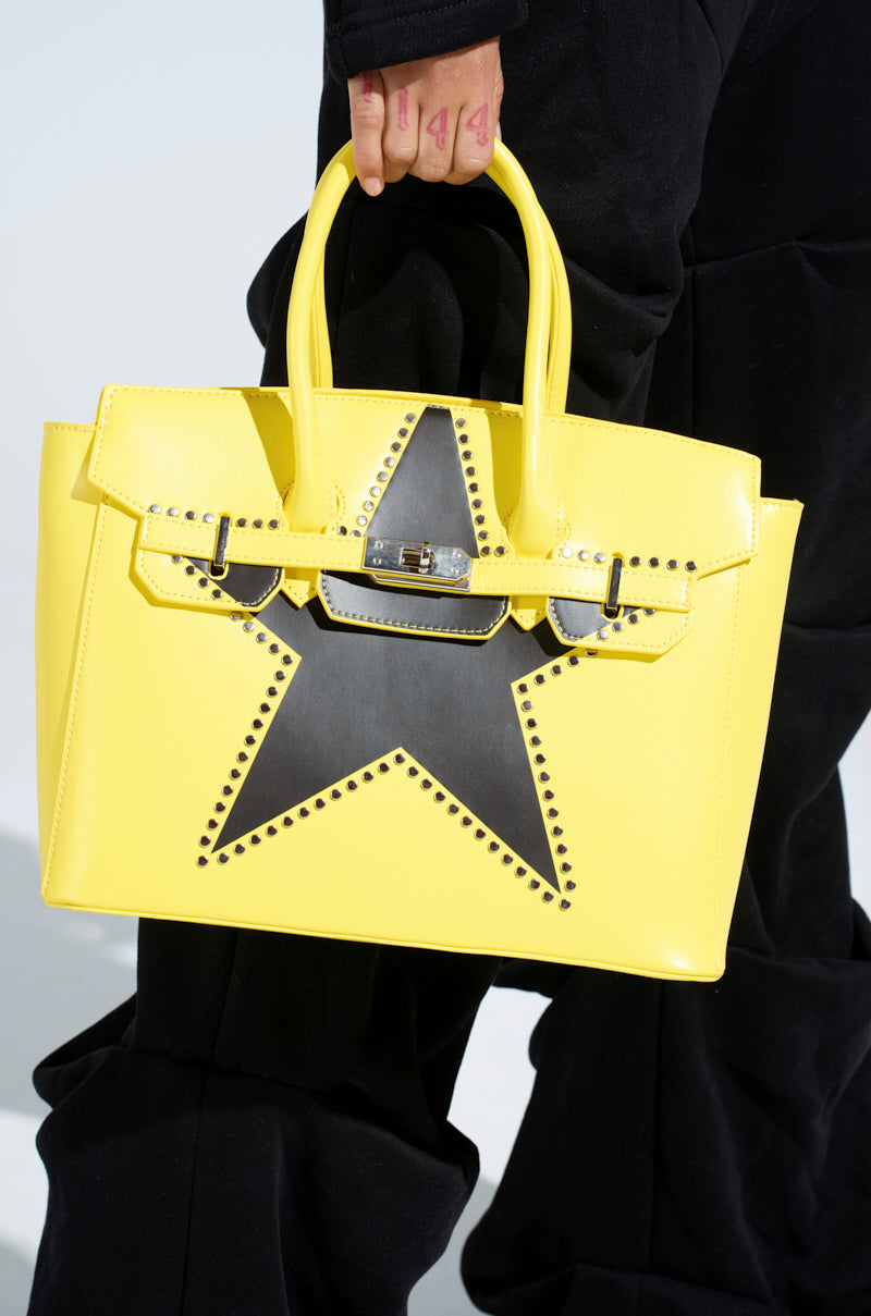 A person holding the STAR POWER TOTE, a bright yellow faux leather bag with a structured shape, black star motif, and silver stud accents, paired with black pants.