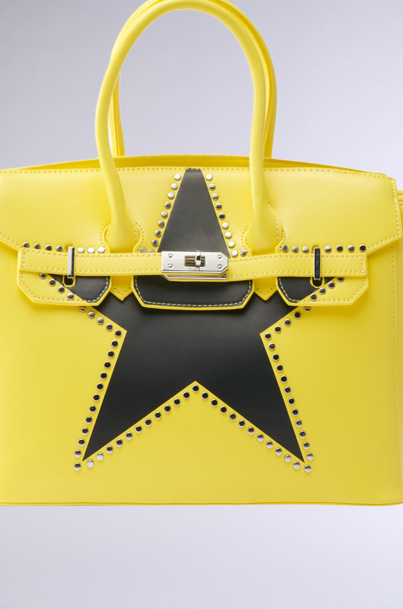 The STAR POWER TOTE is a bright yellow faux leather bag with a bold black star and silver studs on the front, two handles, a silver clasp closure, and a structured silhouette.
