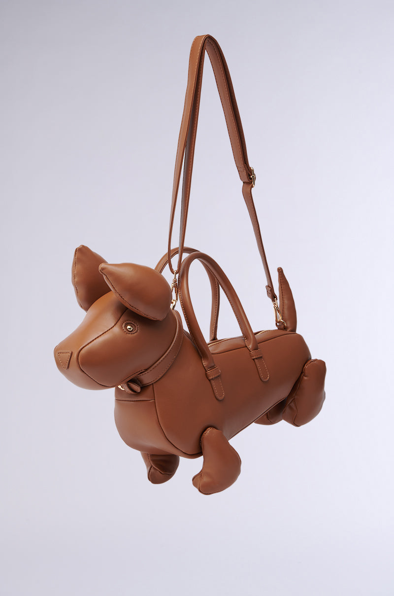 The I'M REALLY A DOG PURSE is a brown faux leather bag shaped like a dachshund, featuring floppy ears, legs, and a tail, and is shown hanging by its long shoulder strap against a simple light background.