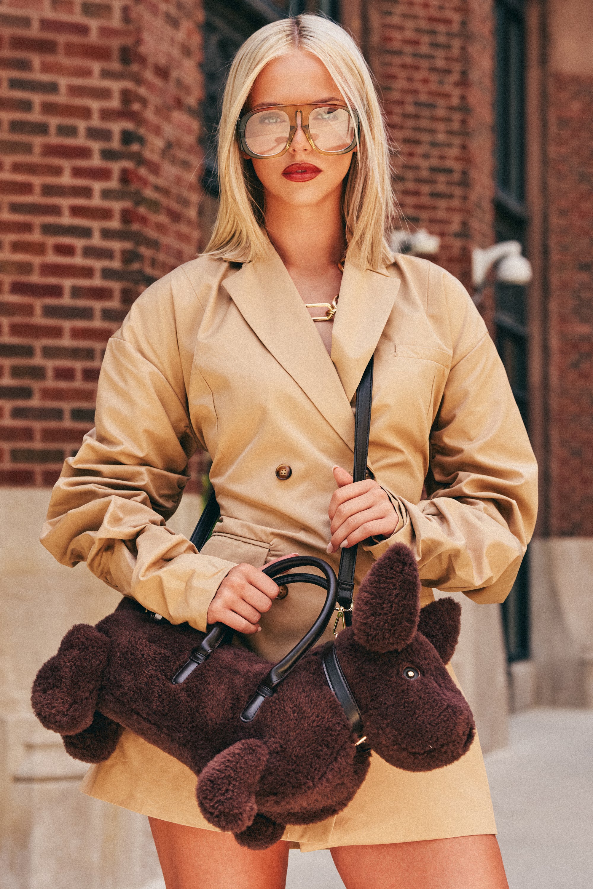 A woman in a tan trench coat and oversized glasses stands on a city sidewalk in front of a brick building, holding the WOOF WOOF PURSE—a novelty streetwear accessory designed to look like a brown faux fur dog.