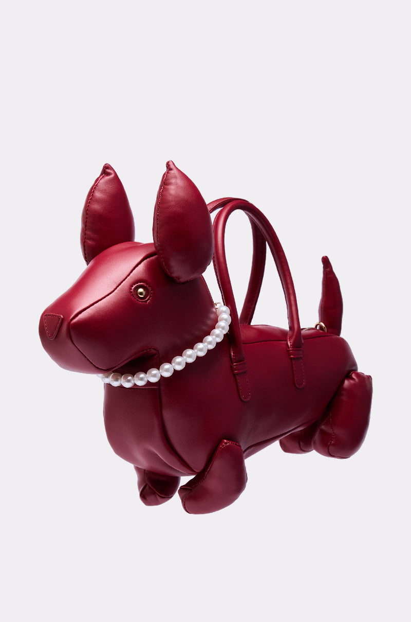 The PRETTY BUT I BITE BACK PURSE is a shiny red faux leather dog-shaped bag with upright ears, a pearl necklace, two handles on its back, and a detachable shoulder strap, displayed against a plain light background.