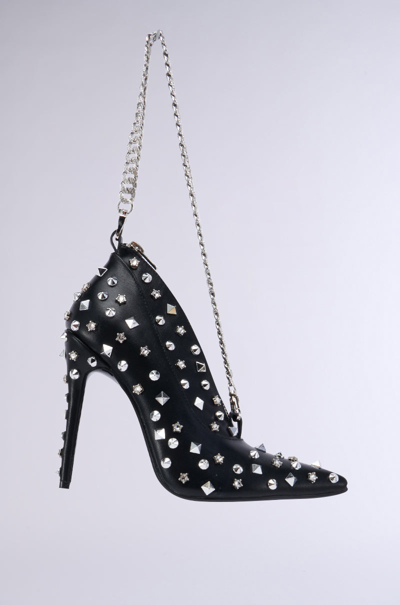 The SUGAR AND SPICE SHOE PURSE is a black high-heeled shoe-shaped purse adorned with an array of silver studs and spikes. Made from faux leather, this accessory features a pointed toe and a thin stiletto heel. A silver chain attached to the back allows for easy carrying or suspension. The item is showcased against a plain, light-colored background.
