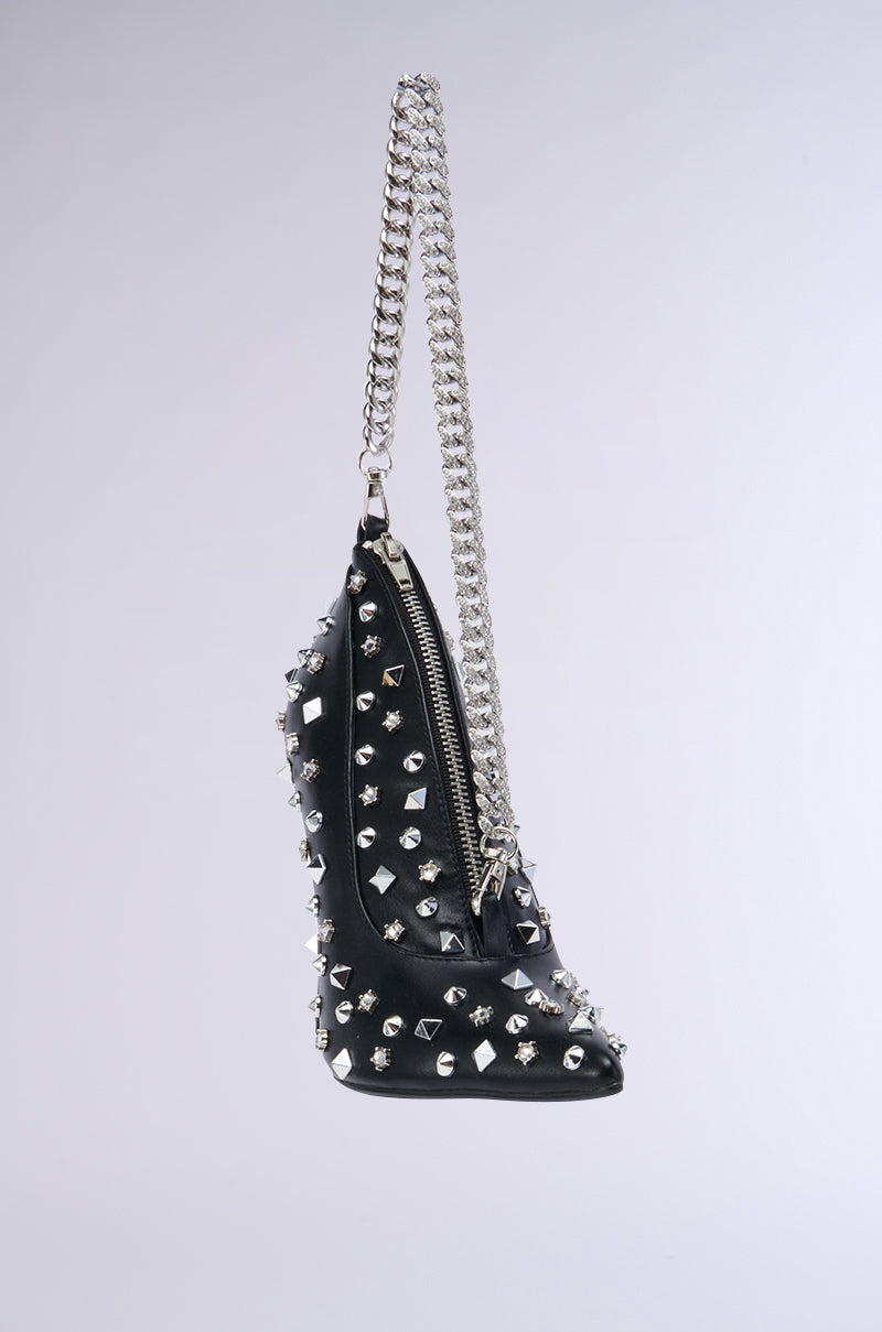 The SUGAR AND SPICE SHOE PURSE, a black faux leather handbag with a silver chain strap, hangs against a light-colored background. It is adorned with metallic silver pyramid studs and features a zippered closure. The design is both edgy and stylish, showcasing a structured, angular shape reminiscent of stiletto heels.