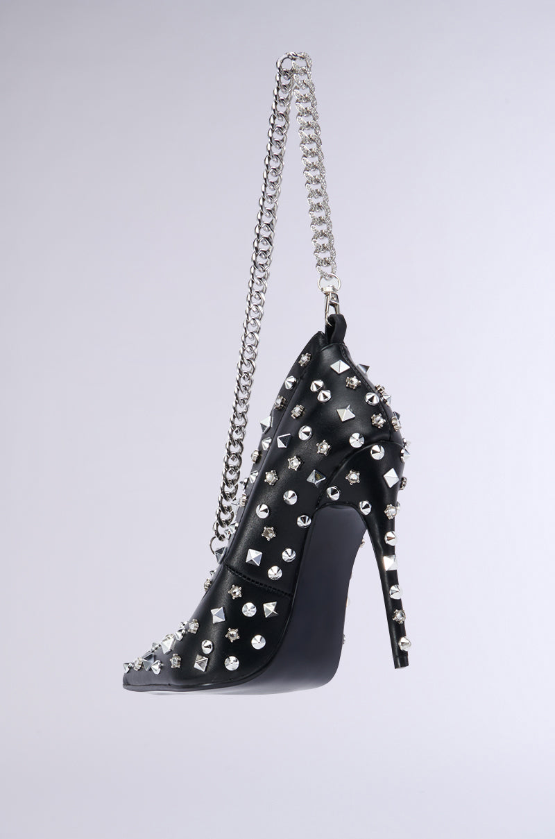 The SUGAR AND SPICE SHOE PURSE, a black high-heeled shoe adorned with silver studs and crystals, is suspended by a silver chain against a plain white background. The purse features a pointed toe and stiletto heel, blending punk rock aesthetics with sophisticated design elements in faux leather.
