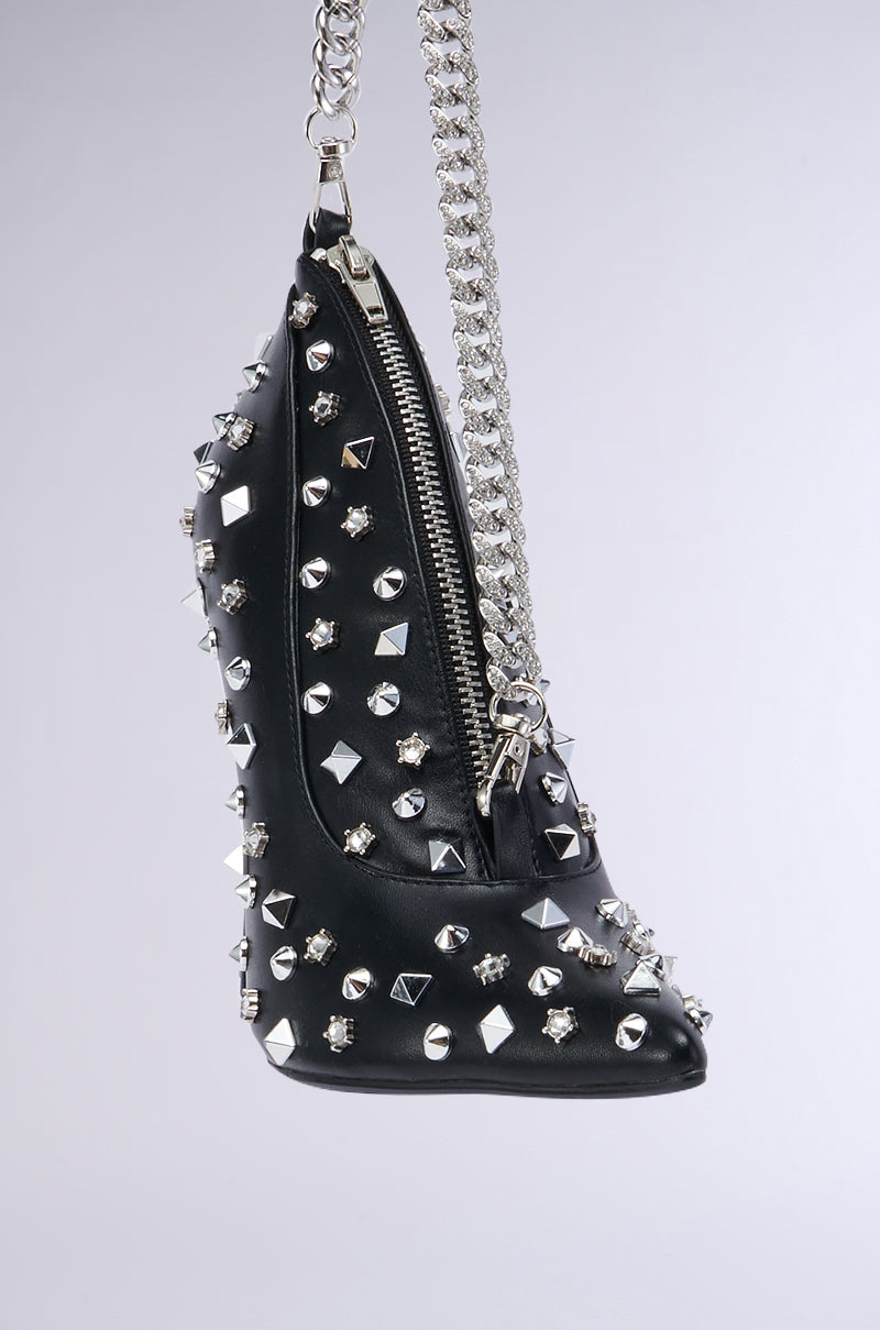The SUGAR AND SPICE SHOE PURSE, a black wedge shoe purse embellished with silver studs of various sizes and a conspicuous side zipper, dangles from a silver chain. This faux leather accessory features an edgy, punk-inspired design with a pointed toe and decorative elements that make it visually striking.
