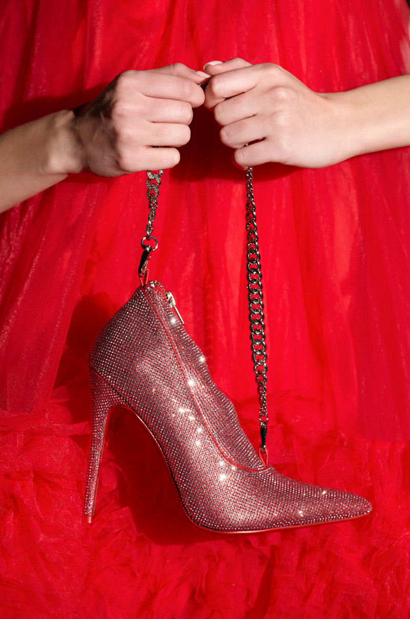 Close-up of hands holding a chain attached to the IN MY RED BOTTOMS PURSE, a glittery red stiletto heel. The person wears a vibrant red, textured garment matching the purse, creating a cohesive and bold fashion statement.