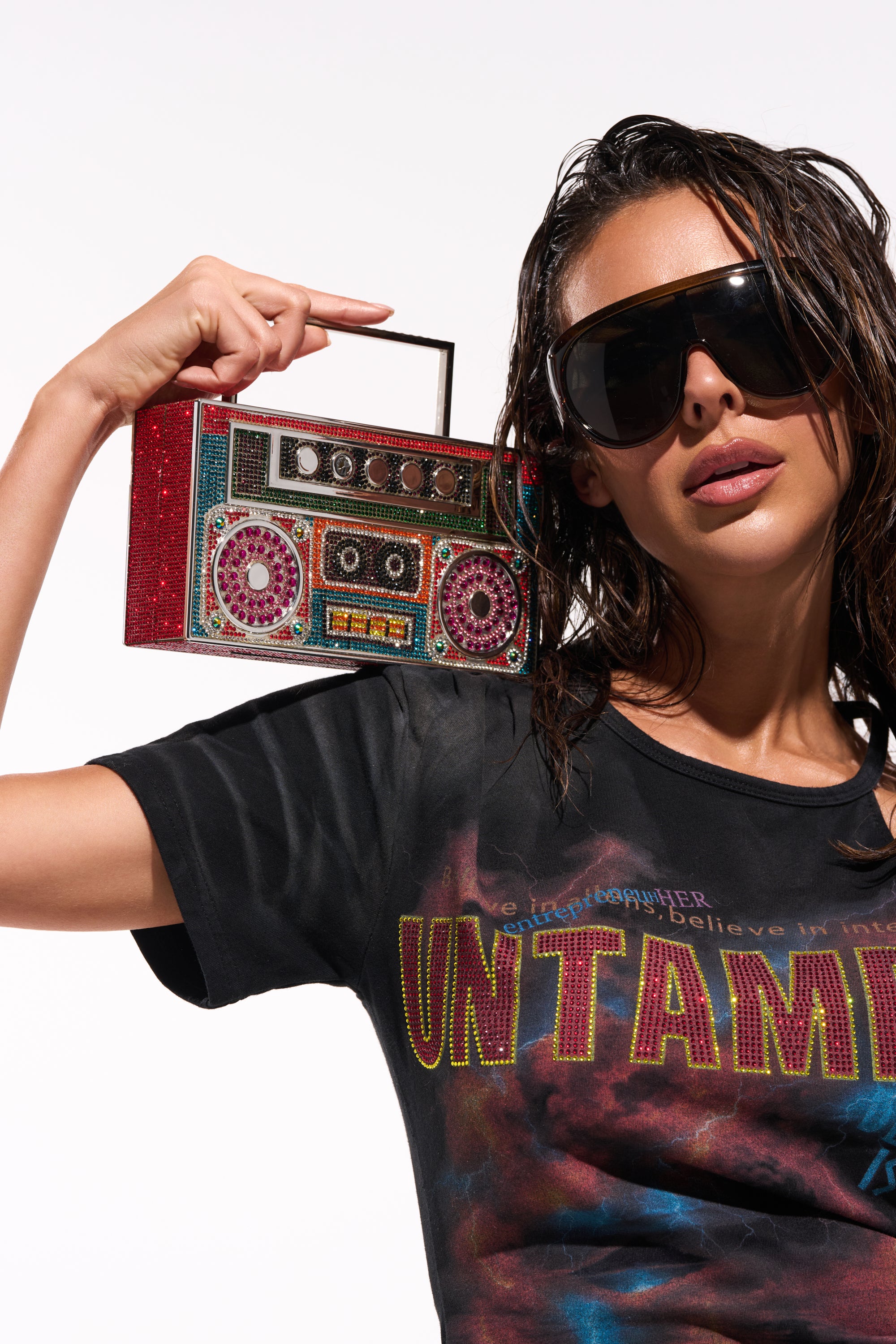 A woman in oversized sunglasses and a black "UNTAMED" tee poses confidently with the TURN ME UP BLING BOOMBOX PURSE on her shoulder, wet wavy hair framing her face, set against a white background.