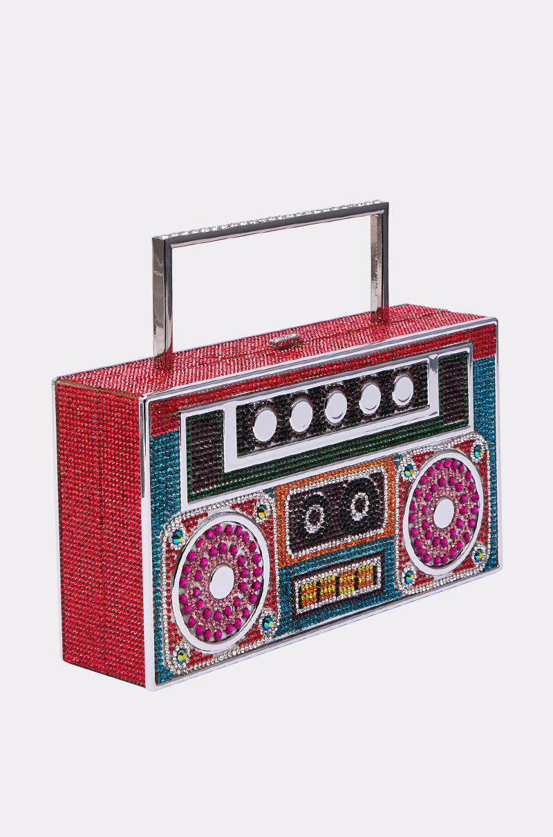 The TURN ME UP BLING BOOMBOX PURSE is a rhinestone bag shaped like a retro boombox, sparkling with red, teal, pink, and yellow stones. It features a sleek rectangular metal handle on top.