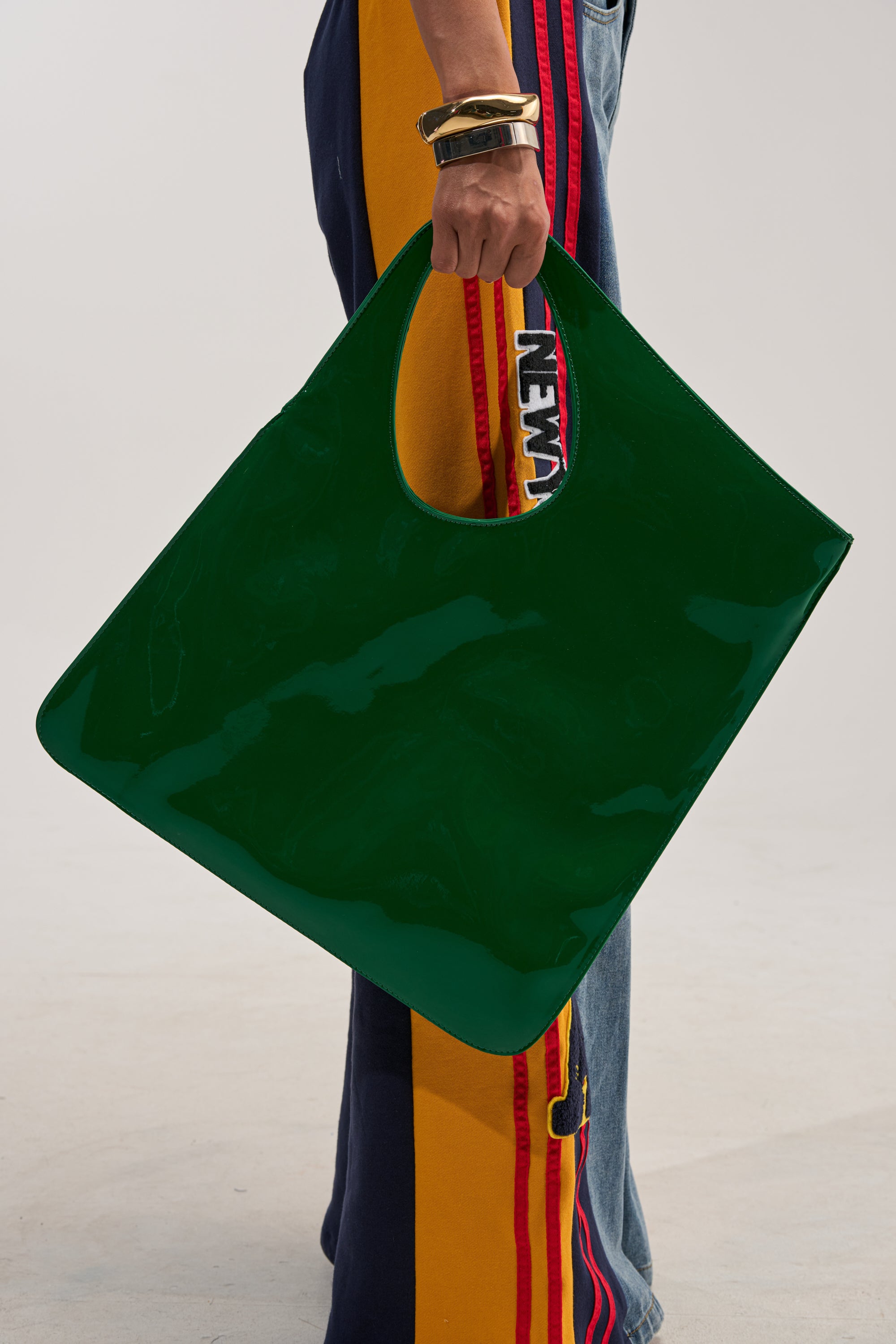 A person dressed in yellow and blue pants with red stripes holds the FEELING ELECTRIC OVERSIZED BAG, a large, glossy dark green patent leather accessory featuring circular handles. Gold bracelets adorn their wrist.