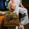 A person in light denim leans against a pool table, holding the CHROME LIFE PURSE—a gold embossed bag with an ornate floral design. Only their lower body and hand are shown; the pool table and balls blur in the background.