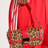 A person dressed in red pants with white stripes down the sides holds an HONEST DEAL WILD ONE CHEETAH MINI PURSE with faux pony hair, featuring red accents and a prominent metallic clasp on the front. The handbag also includes a detachable shoulder strap for added versatility.