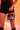 A person in a black outfit with pink floral details holds the JACKY BELT BAG, a small structured frame bag with short handles. The faux leather accessory pops against the orange background, adding warmth and vibrancy to the scene.