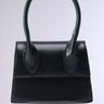 The JACKY BELT BAG is a small, elegant accessory in black faux leather with a rectangular shape and structured design. It features two short, rounded handles emerging from the top and minimalistic stitching details. The bag has a smooth, polished finish against a plain light background and includes a removable shoulder strap.