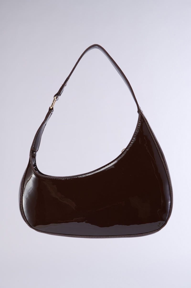 The CHOCOLATE DREAMS PU PURSE is a glossy, dark brown handbag with a sleek, minimalistic design. It features a single shoulder strap adorned with gold-tone hardware and boasts an asymmetrical silhouette. The bag's curved, semi-rectangular shape creates a modern, stylish look. 0