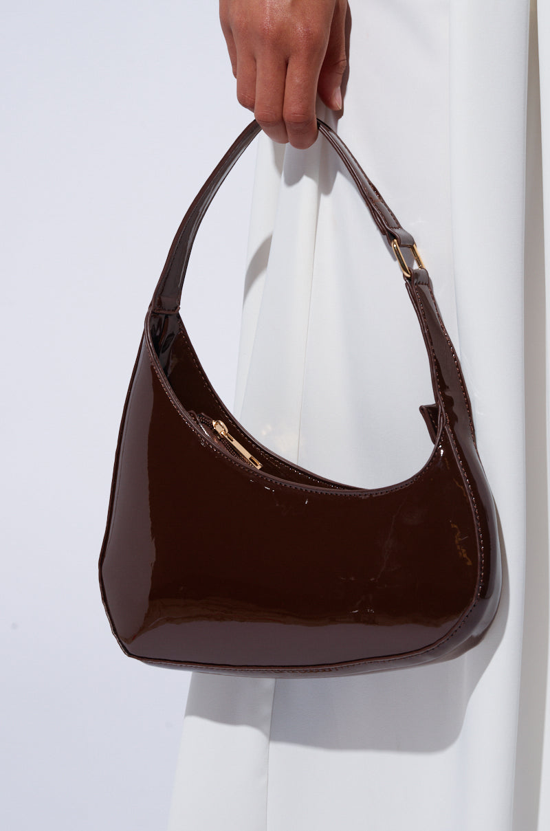Someone is holding the CHOCOLATE DREAMS PU PURSE, a petite, dark brown handbag made of faux patent leather with a shiny finish. The purse features a short strap and zipper closure that catches the light. The person is dressed in white clothing. 1