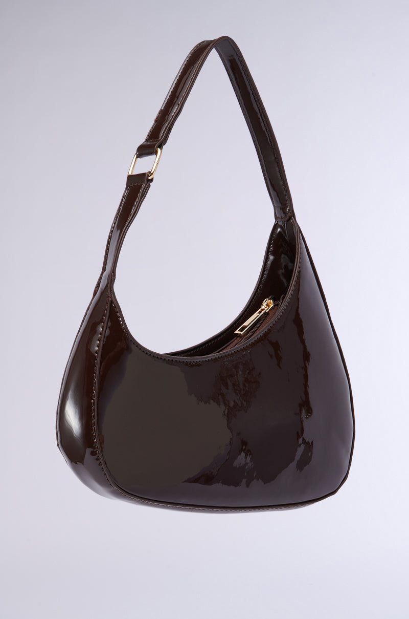 A CHOCOLATE DREAMS PU PURSE with a crescent shape is pictured against a plain background. Its asymmetrical silhouette features a curved handle with a gold buckle and a top zipper closure, partially open to reveal a gold zipper pull. The faux patent leather surface is shiny and reflective. 2