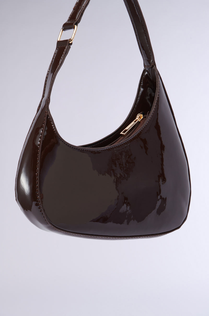 The CHOCOLATE DREAMS PU PURSE is a sleek, small shoulder bag crafted from shiny black faux patent leather. It features a gold-tone zipper on the front and a single strap with a gold buckle. The bag has an asymmetrical silhouette and is photographed against a plain gray background. 3