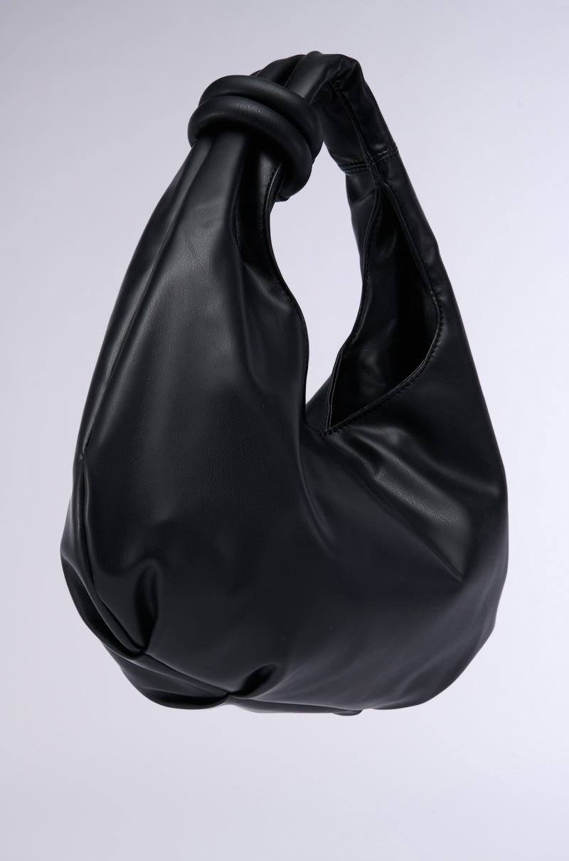 The TOO TRENDY PU TOTE from the AKIRA Label is showcased against a simple gray background. This black faux leather handbag features a distinct design with a single knotted handle at the top and a slouchy, rounded body, giving it a modern and stylish look.