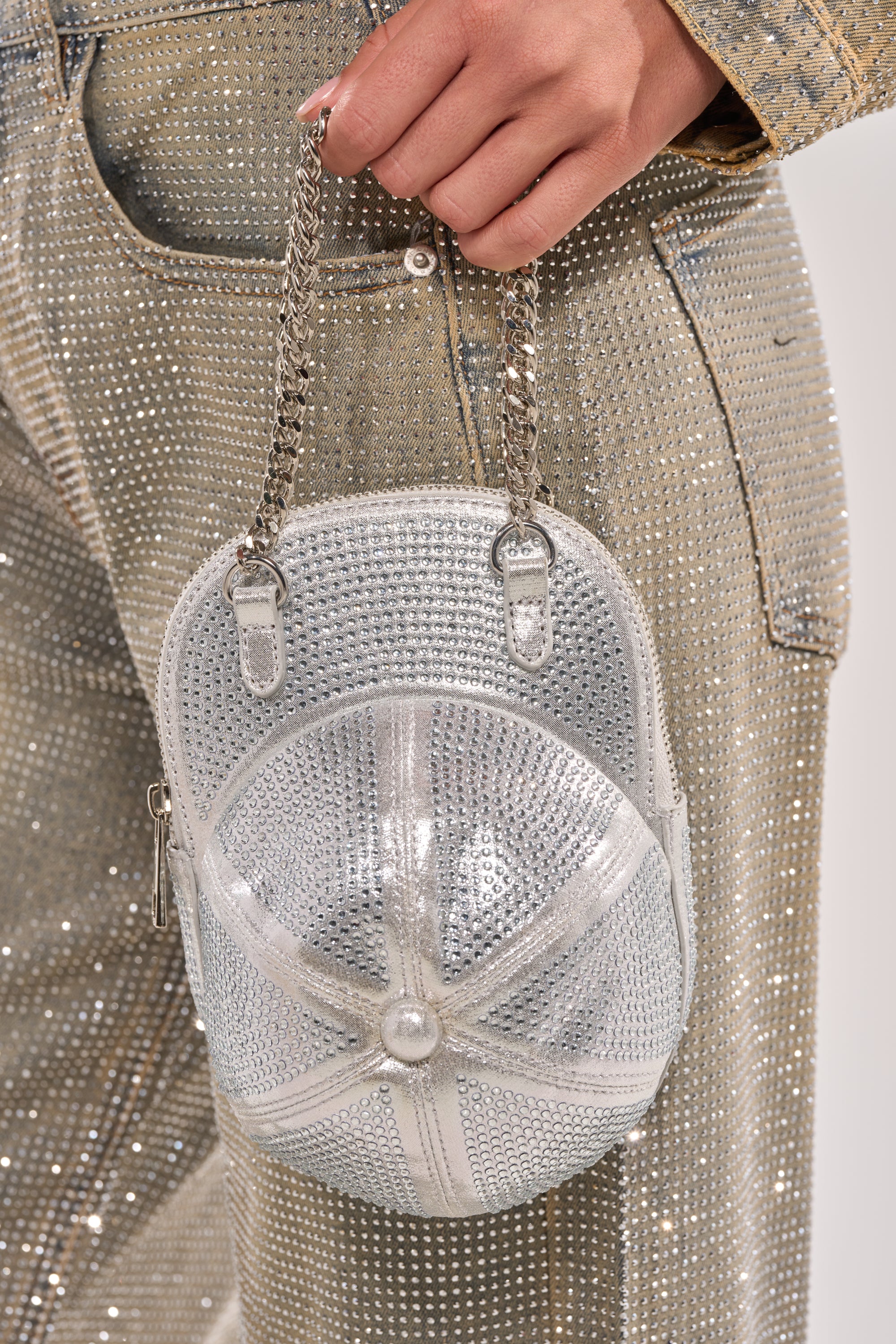 A person holds the NO CAP BLING PURSE, a metallic silver hat-shaped bag encrusted with rhinestones and featuring a chain handle, while wearing sparkling, rhinestone-studded gold pants. 1