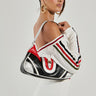 A woman models a chic white off-the-shoulder ensemble adorned with red and black stripes, while carrying the SPORT MODE PURSE, a faux leather designer handbag featuring a varsity-inspired design. Her hair is elegantly styled in an updo, complemented by large hoop earrings, all set against a plain white background.