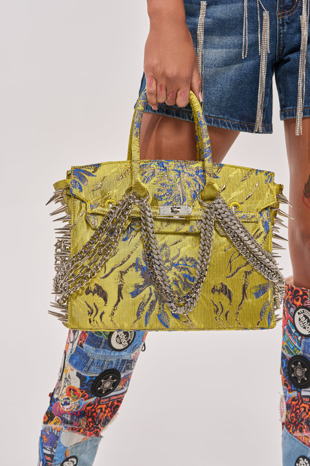 A person in denim shorts holds the LETS GO EMBELLISHED TOTE, featuring metal spikes and silver chains. Their boots have colorful patches, and the background is plain white.
