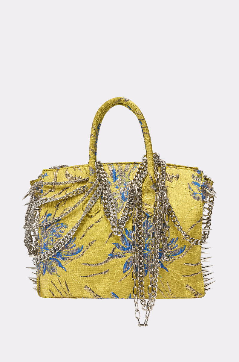 The LETS GO EMBELLISHED TOTE is a chartreuse floral brocade handbag with blue patterns, silver spikes, and layered chains draped over the handles and front—a striking spring statement piece.