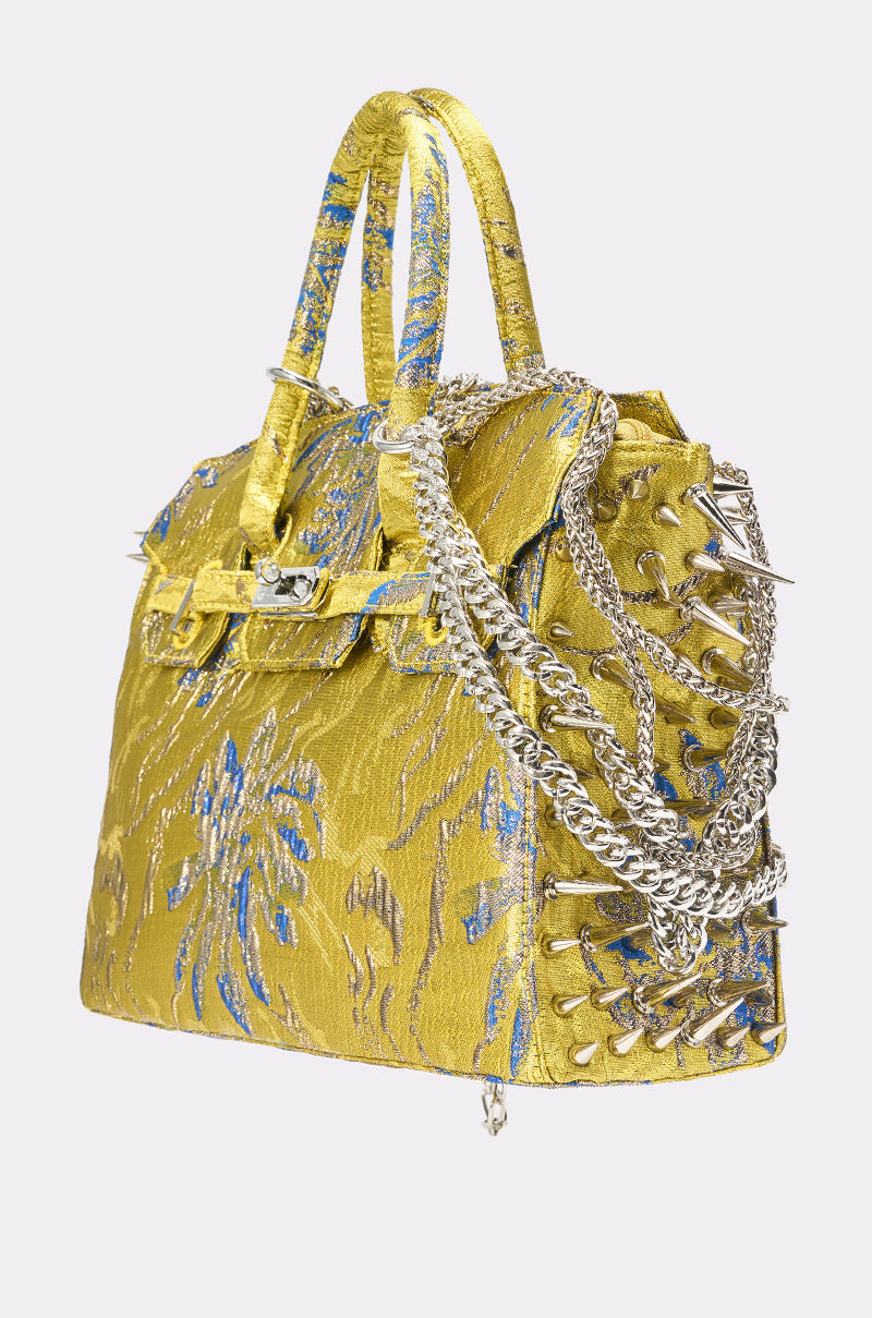 The LETS GO EMBELLISHED TOTE in gold and blue features patterned details, lace-up sides, dual handles, and metallic hardware, making a bold statement against a plain light background.