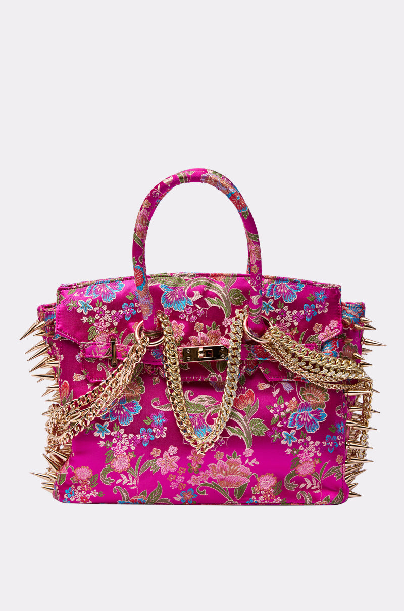 The CECE SPIKE TOTE is a bold, rectangular tote in vibrant fuchsia with a colorful floral pattern, gold chain handles, and large gold spikes accenting the sides and edges.