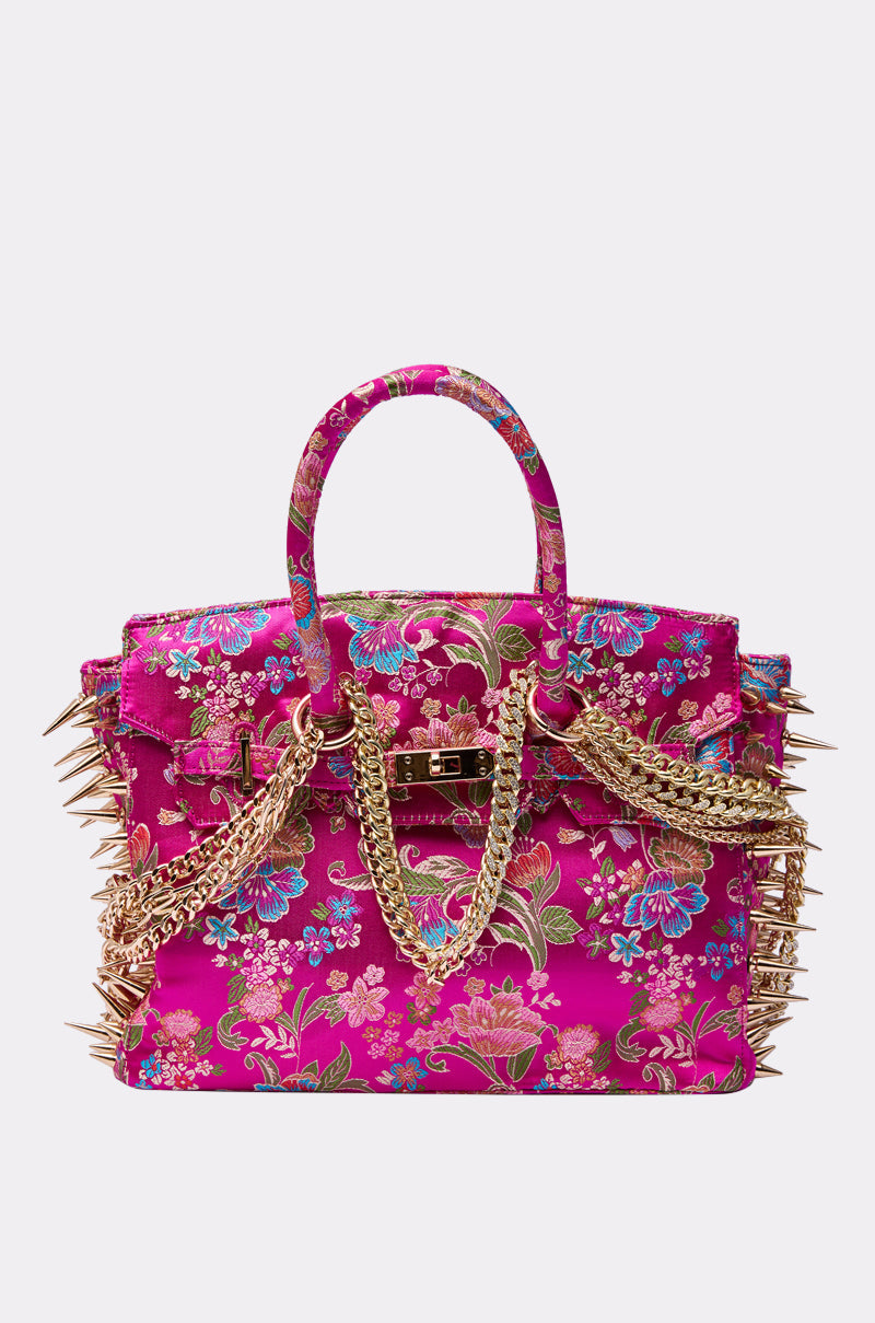 The CECE SPIKE TOTE is a bold, rectangular tote in vibrant fuchsia with a colorful floral pattern, gold chain handles, and large gold spikes accenting the sides and edges.
