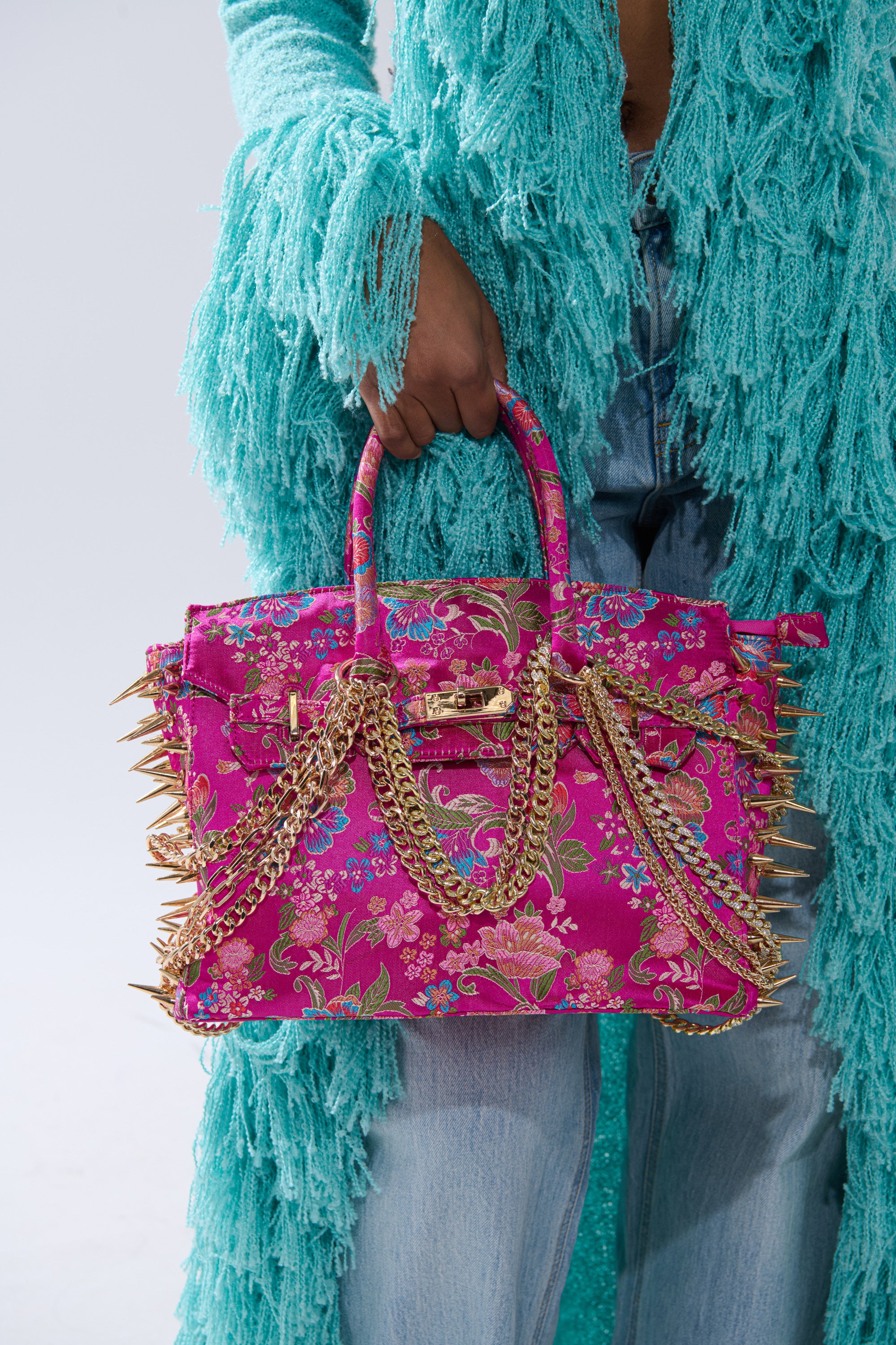 A person in a turquoise fringed jacket and blue jeans holds the CECE SPIKE TOTE—a fuchsia floral bag with gold spikes, chains, and embellishments.