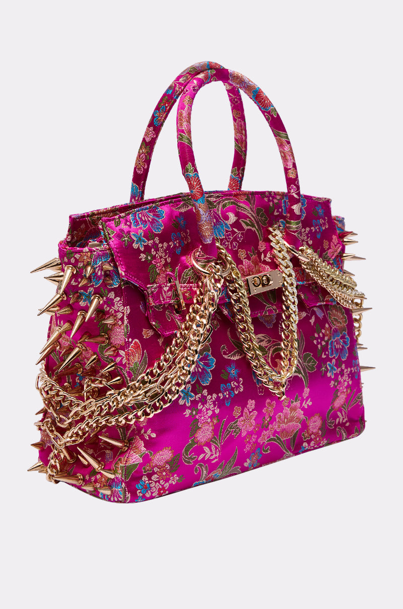 The CECE SPIKE TOTE is a vibrant fuchsia floral tote with gold spikes, decorative gold chains, two handles, and an ornate brocade pattern—making it a striking statement accessory.