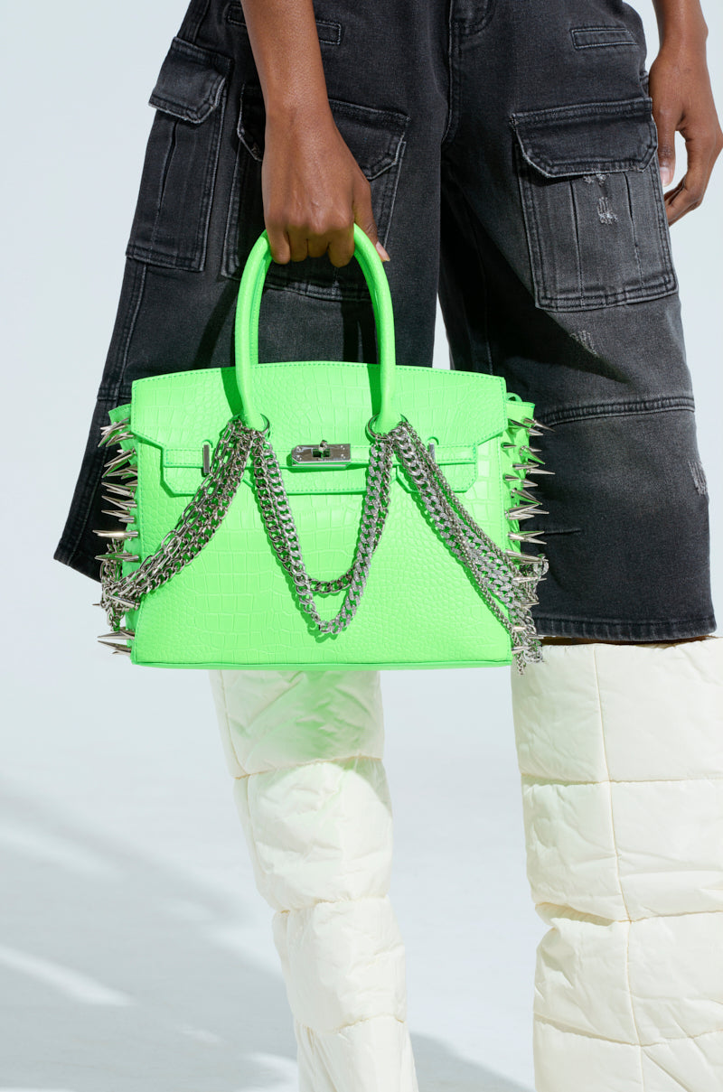 A person carrying the COME SLIDE EMBELLISHED TOTE, a neon green handbag embellished with silver chains and spikes, is dressed in black cargo shorts with white quilted pants underneath. The faux crocodile leather bag stands out against the light neutral background.
