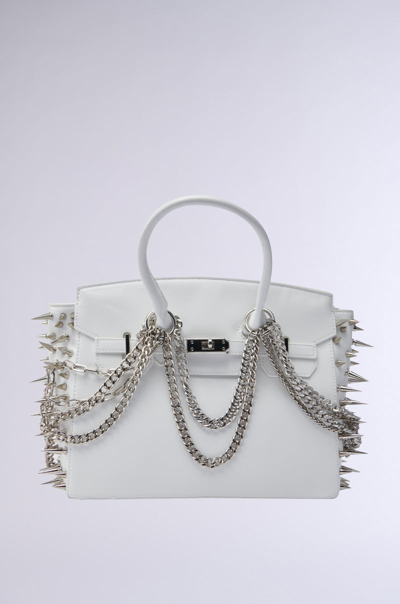 The STAY BLESSED SPIKE PURSE is a white faux leather handbag with silver spikes on the sides and silver chains draped over the front handles and body, set against a light gray backdrop.