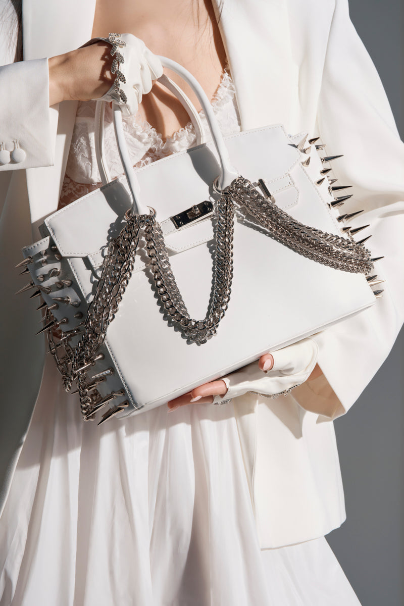 A person in a white suit and lace dress carries the STAY BLESSED SPIKE PURSE, a fashion-forward faux leather bag featuring silver spikes and metal chains for a bold, edgy look.