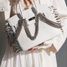 A person in a white suit and lace dress carries the STAY BLESSED SPIKE PURSE, a fashion-forward faux leather bag featuring silver spikes and metal chains for a bold, edgy look.