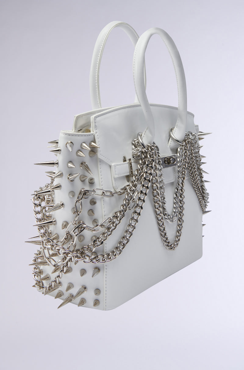 The STAY BLESSED SPIKE PURSE is a fashion-forward white faux leather handbag, decorated with large silver spikes and multiple silver chains, displayed against a plain light gray background.