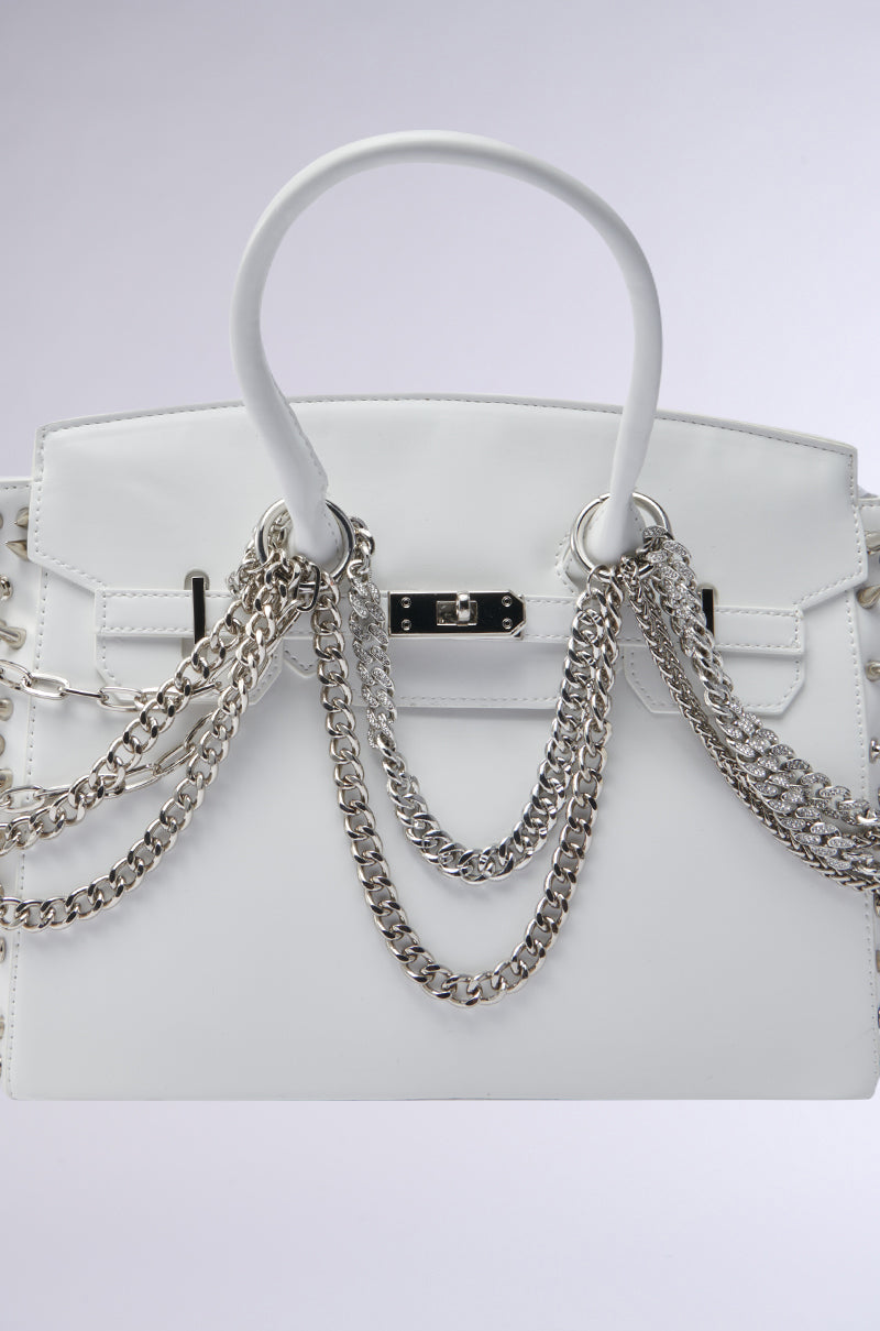 The STAY BLESSED SPIKE PURSE is a white faux leather handbag with silver chain straps, a silver clasp and handle, set against a light gray background—a chic addition to any modern wardrobe.