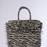 The TRY ME EMBELLISHED TOTE, a zebra print handbag with two handles and bold metallic spike accents along the edges and top, is displayed against a plain light background.