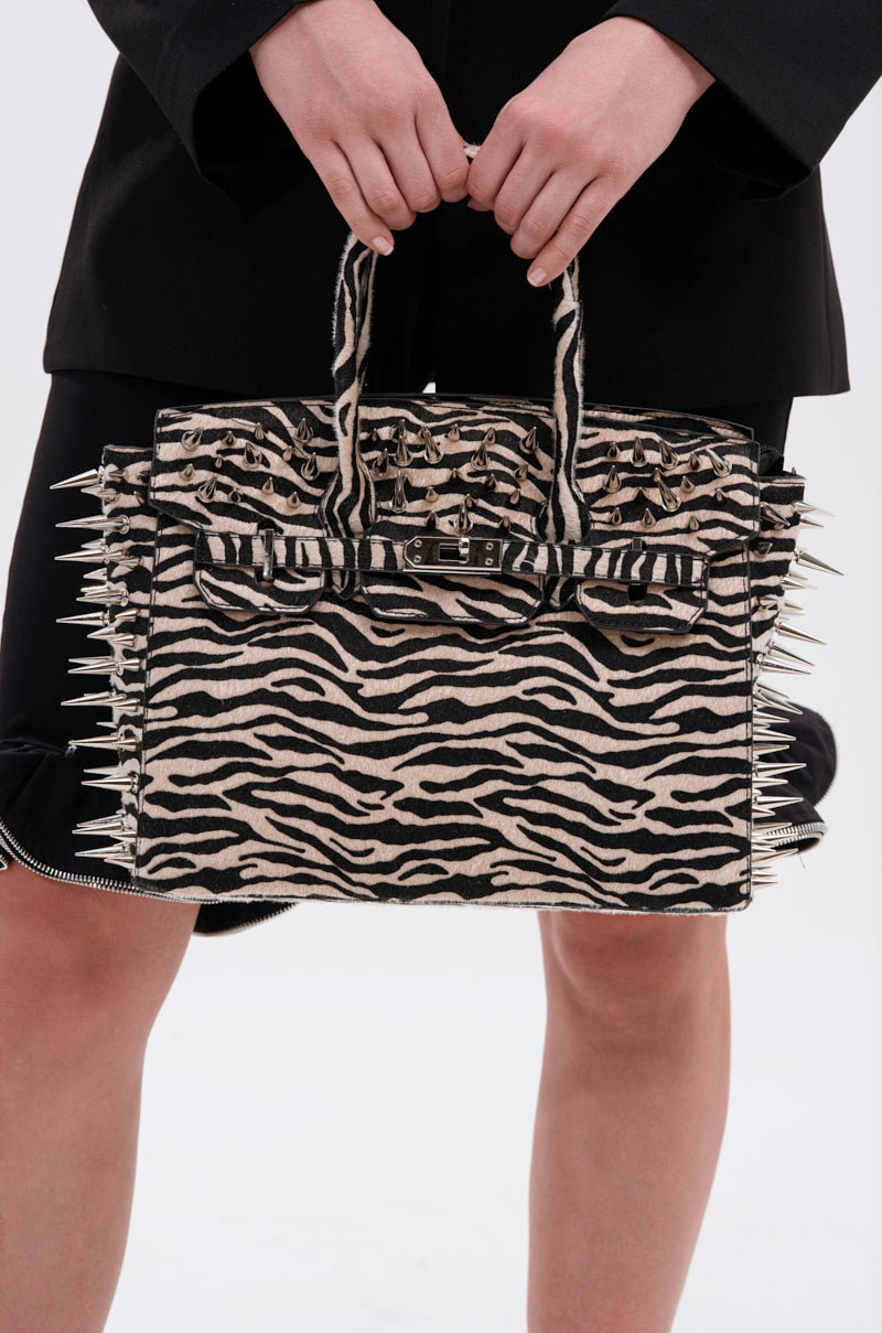A person holds the TRY ME EMBELLISHED TOTE featuring zebra print, metallic spikes, and stud details, paired with a black outfit; focus is on the bag and hands against a plain background.