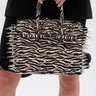 A person holds the TRY ME EMBELLISHED TOTE featuring zebra print, metallic spikes, and stud details, paired with a black outfit; focus is on the bag and hands against a plain background.