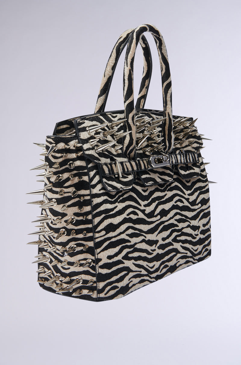 The TRY ME EMBELLISHED TOTE is a faux fur handbag with bold black and white zebra stripes, accented by metallic spikes for a standout, punk-inspired style.