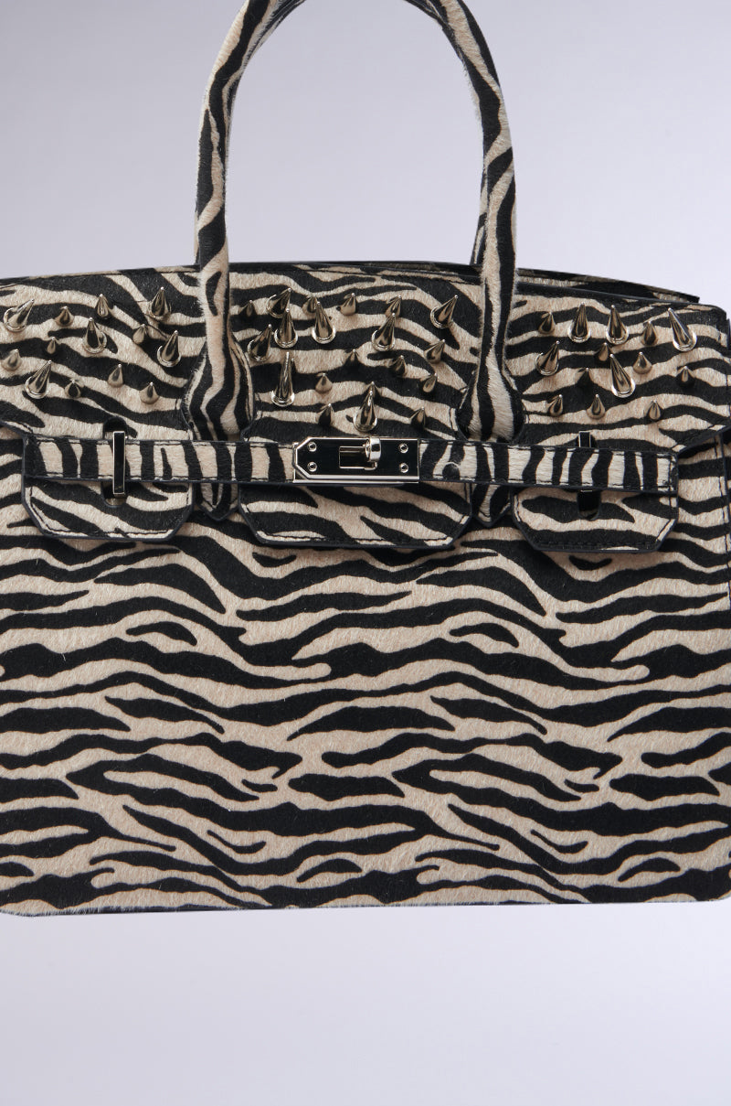 The TRY ME EMBELLISHED TOTE features a zebra print, bold metallic spikes on the top and handles, and a decorative front clasp—perfect for making a striking statement.