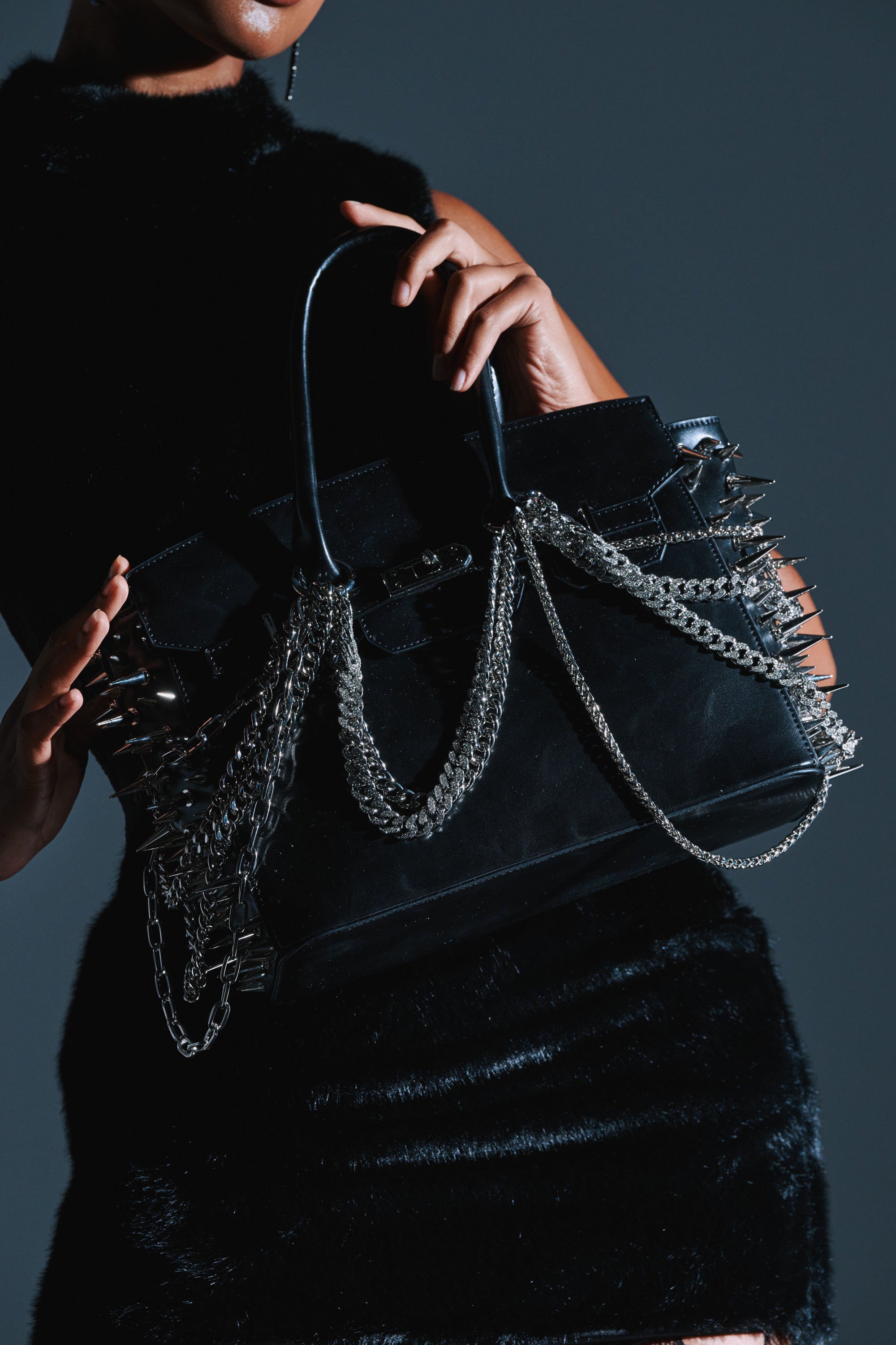 A person in a black fuzzy dress holds the I BITE BACK EMBELLISHED TOTE, a faux leather handbag with silver spike embellishments and chains, set against a dark background. 2