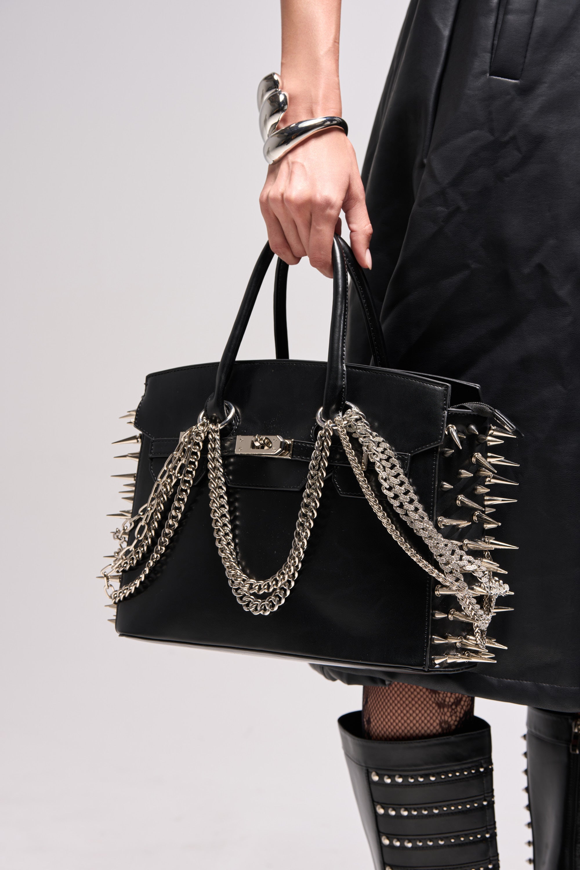 A person in black leather attire holds the I BITE BACK EMBELLISHED TOTE, featuring silver spikes and chains. They pair it with a silver bracelet, fishnet stockings, and studded black boots against a plain light background. 1