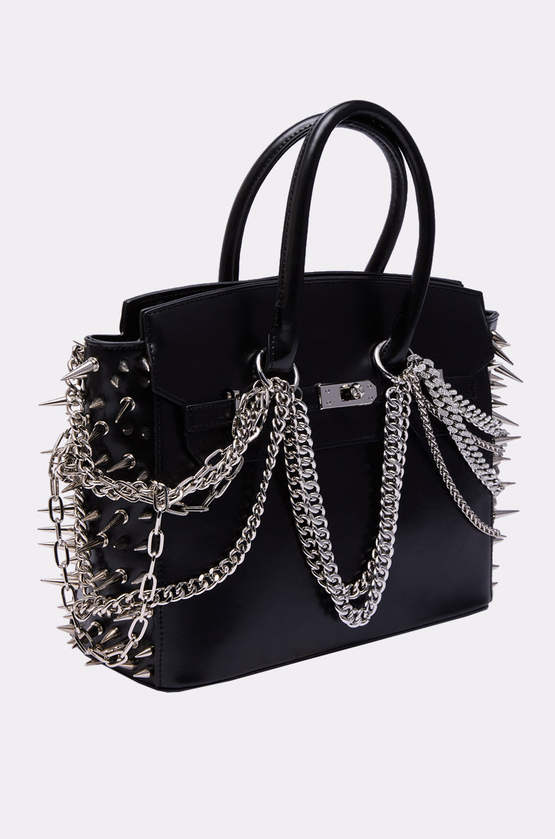 The I BITE BACK EMBELLISHED TOTE is a black faux leather handbag featuring silver spike accents on the sides and layered silver chains across the front and handles, shown against a plain light background. 4
