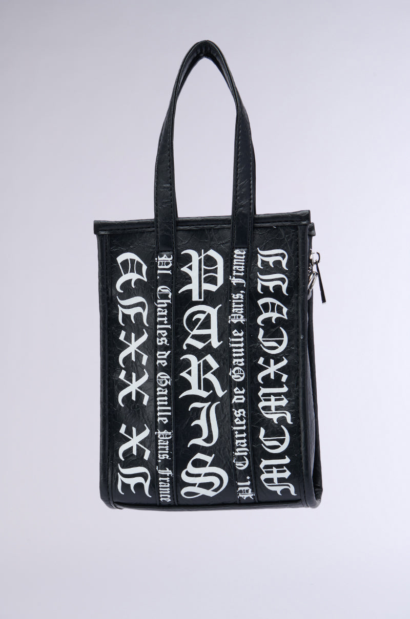 The CHAMPS GRAFFITI PURSE is a black, rectangular tote bag made of faux leather with sturdy handles. The front features bold, white, gothic-style text that reads "PARIS" and various Roman numerals. The bag also has "Charles de Gaulle Étoile Paris" written on the sides in smaller gothic-style text, complemented by subtle graffiti patterning. 1