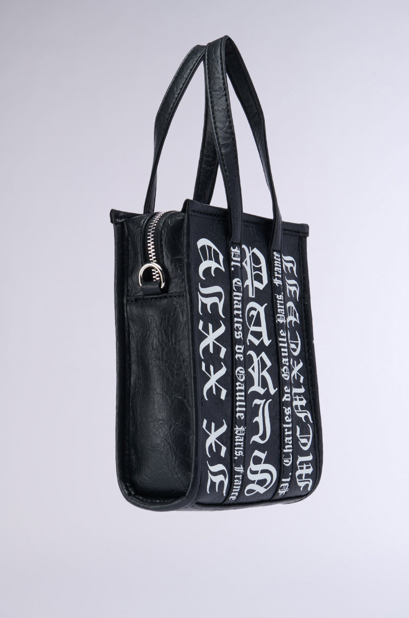 The CHAMPS GRAFFITI PURSE is a black faux leather tote with dual handles, adorned with stylish white Gothic script text on the front and sides. It features a chic silver zipper closure on top, set against a plain and neutral background. 2