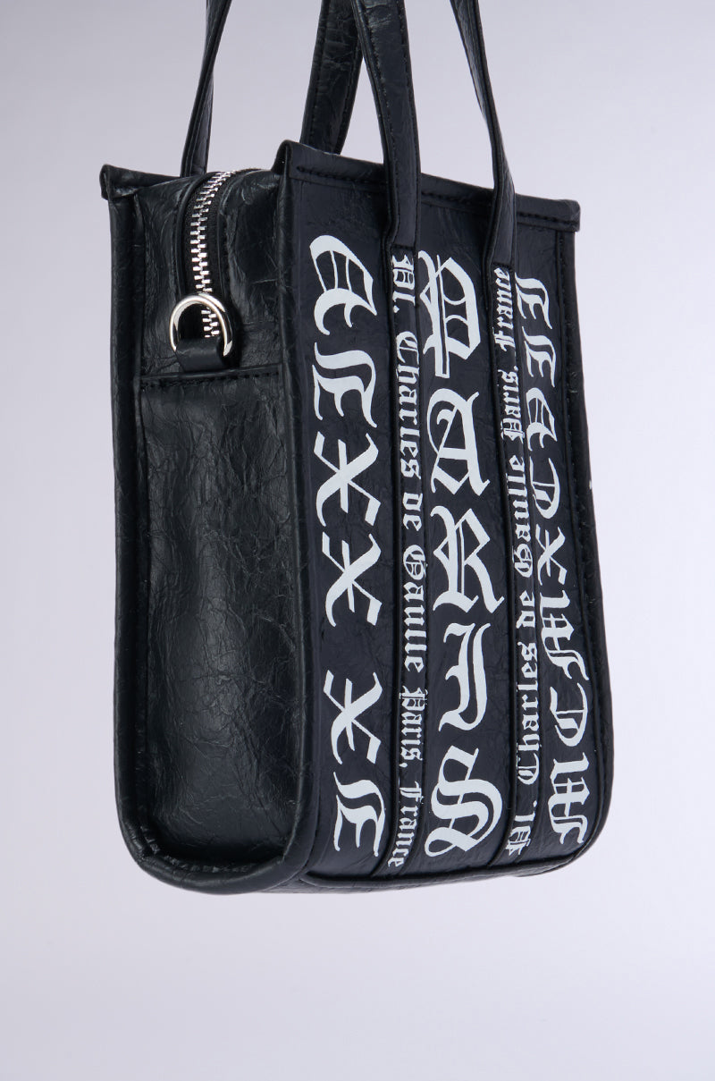 Meet the CHAMPS GRAFFITI PURSE – a chic handbag crafted from black faux leather, adorned with Gothic-style text reading "PARIS" in bold, stylized white letters with a subtle graffiti pattern on the front and straps. It features a side zipper pocket with a metallic zipper pull against a plain, light-colored backdrop. 3