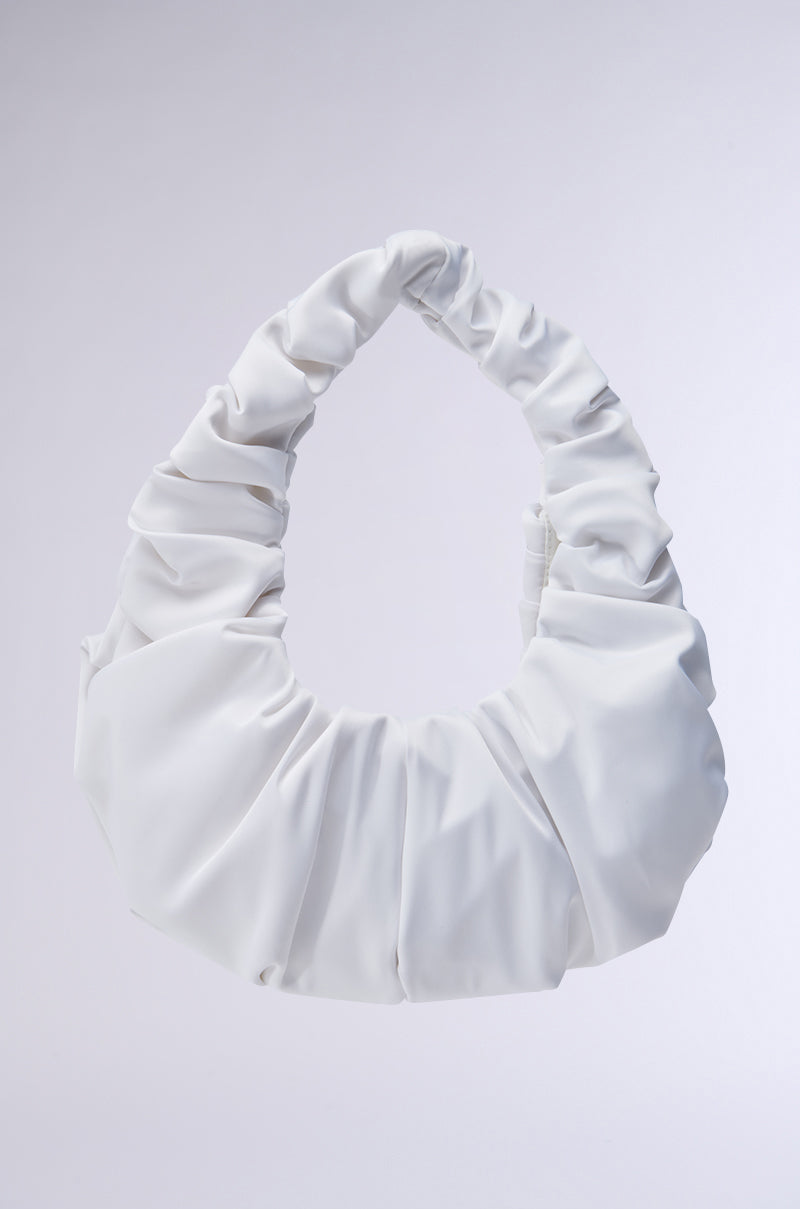 Displayed against a light gray background is the TRU SATIN RUCHED PURSE, a stylish white accessory featuring a ruched design and rounded handle. The smooth, slightly shiny satin material adds a modern and chic touch to its appearance.